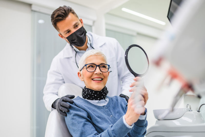 an image of an oral surgeon smiling with a patient into a mirror after she has learned all the factors that can affect the cost of her dental implants 