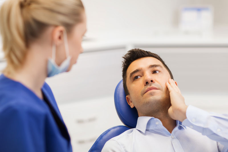 a patient touching the side of his face where he has wisdom tooth pain as he talks to an oral surgeon standing over him about getting a wisdom tooth removal.